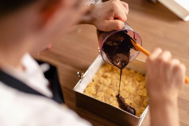 An image of a baker pouring melted chocolate into a pan of baked treats