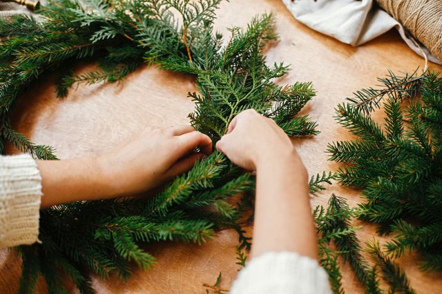 An image of hands holding fir branches, and pine cones, thread, berries, and scissors on wooden table