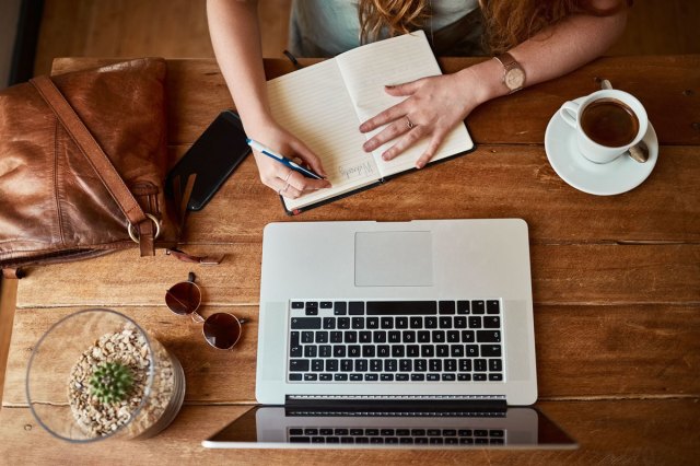 An image of a woman sitting at a wooden table with a laptop and writing in a notebook