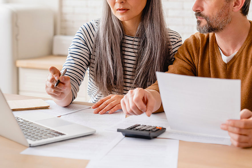 An image of a woman and a man sitting at a desk with a computer, paper, and calculator