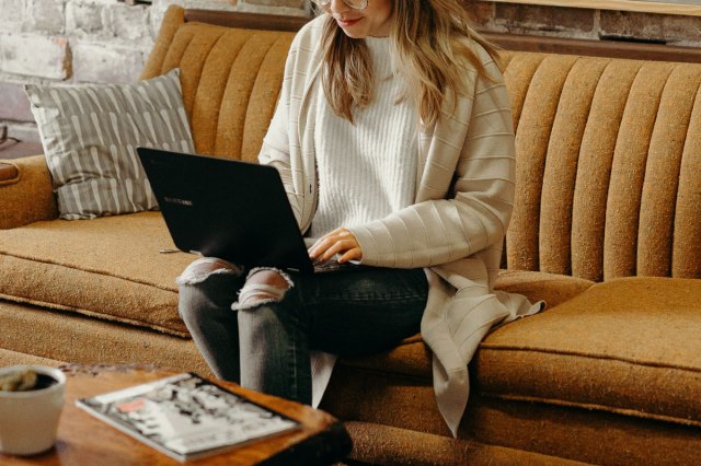 An image of a woman sitting on a couch and typing on a laptop