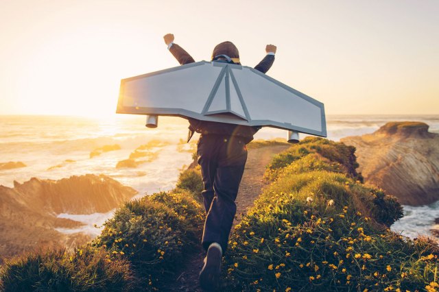 An image of a young boy wearing a homemade jetpack and raising his arms in the afternoon sun while running to take off into the air on an outcropping above the surf