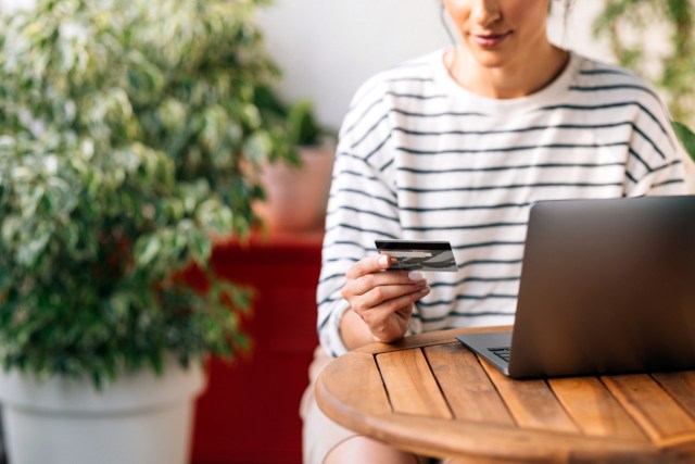 An image of a woman sitting at a table with a laptop and looking at her credit card