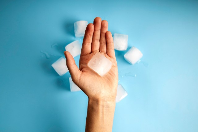 An image of a hand holding an ice cube against a blue background with ice cubes