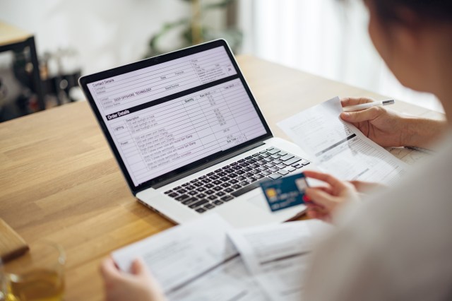 An image of a person holding a credit card an sitting at a table with a laptop and paperwork