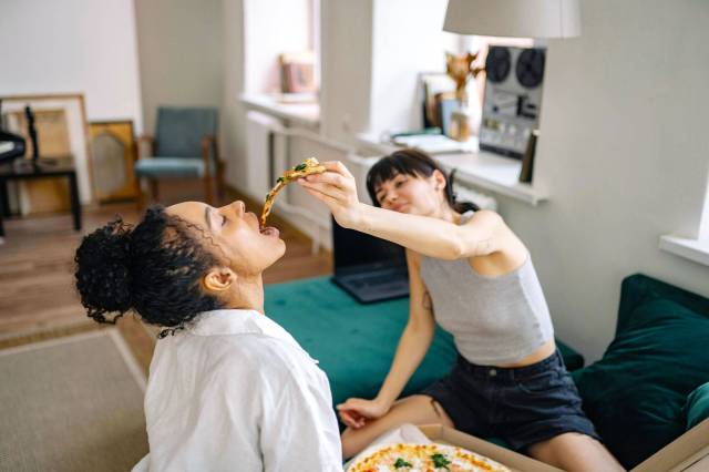 Woman feeding a slice of pizza to another woman on the couch