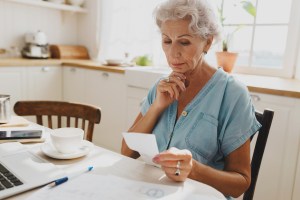 Puzzled worried serious elderly lady accountant with gray hair working from home, examining pay check sitting at kitchen table at home, counting expanses and income. Grandma holding utility bill